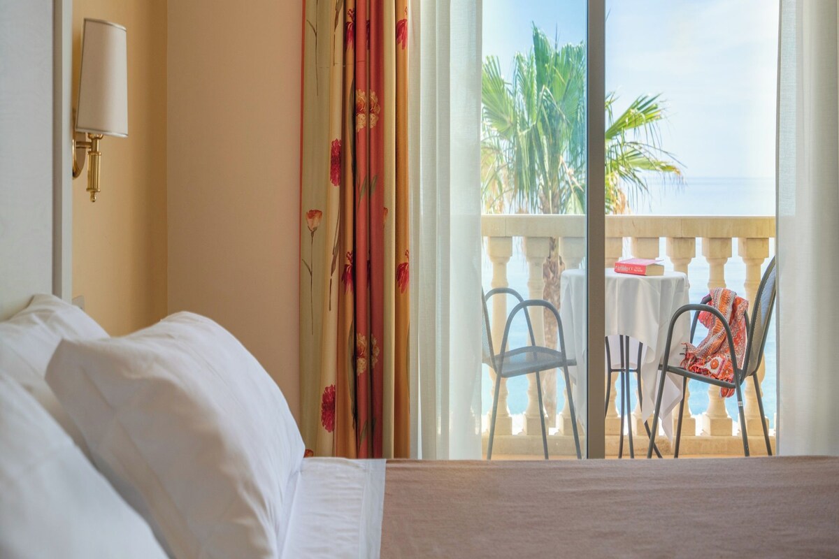 A hotel room with a bed, a nightstand, a lamp, a balcony with a table and chairs, and a view of palm trees and the sea.