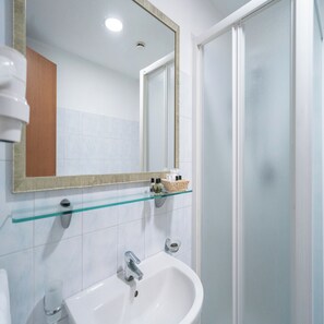 A bathroom with a white sink, a glass shelf with toiletries, a mirror, and a shower door. - Hotel Orizzonte (Letojanni)