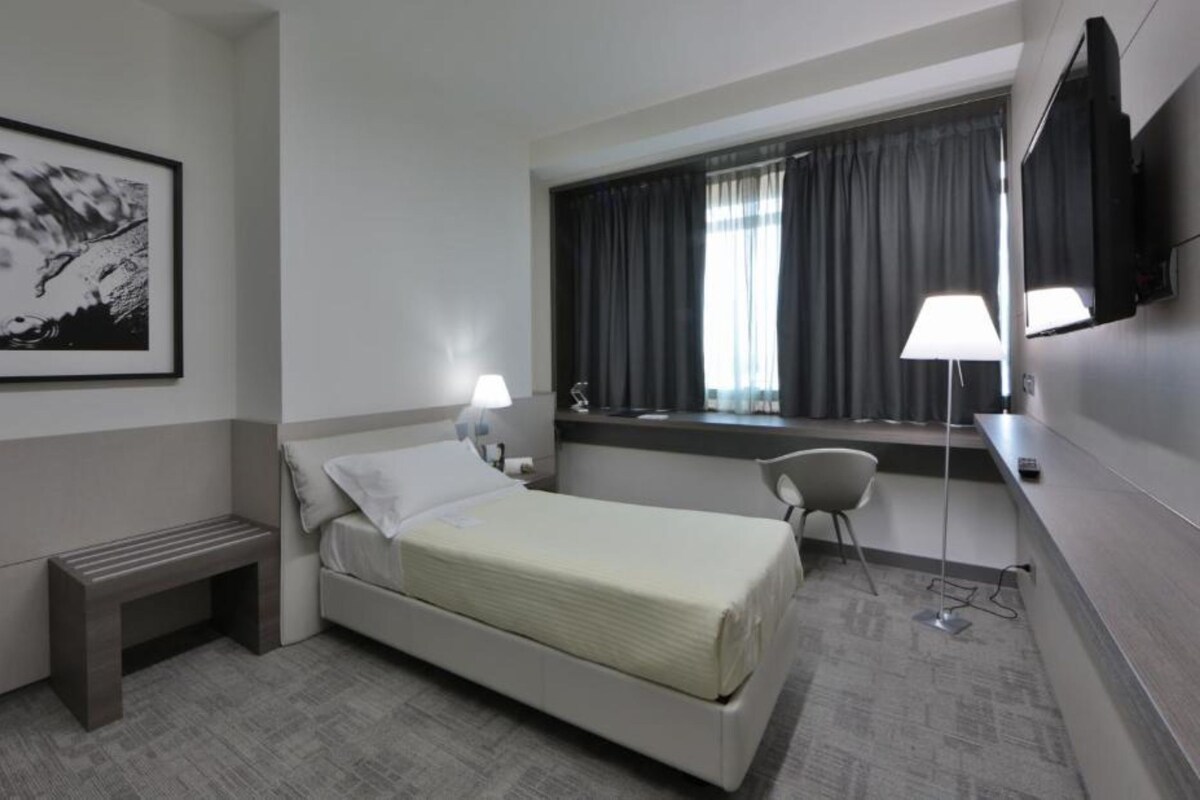 Una habitación de hotel moderna con una cama individual, una mesita de noche, un escritorio con silla, un televisor de pantalla plana, una ventana con cortinas y un cuadro en la pared.