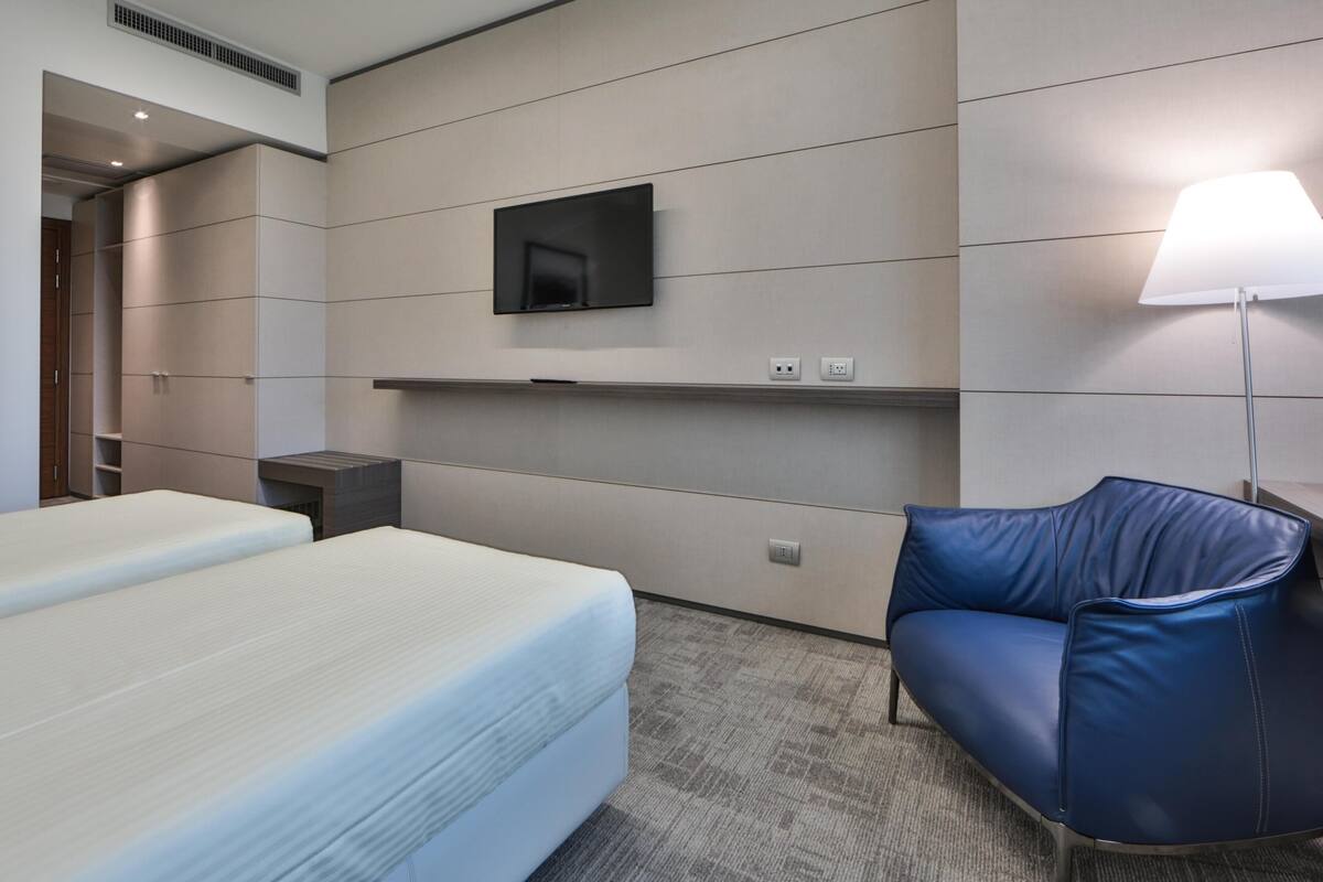 Une chambre d’hôtel avec un lit, un téléviseur fixé au mur, un bureau et une chaise bleue.