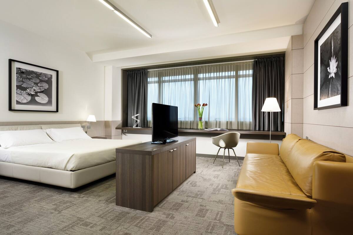 Une chambre d’hôtel avec un lit, un téléviseur posé sur une console en bois, un canapé, un bureau et une grande fenêtre avec des rideaux.