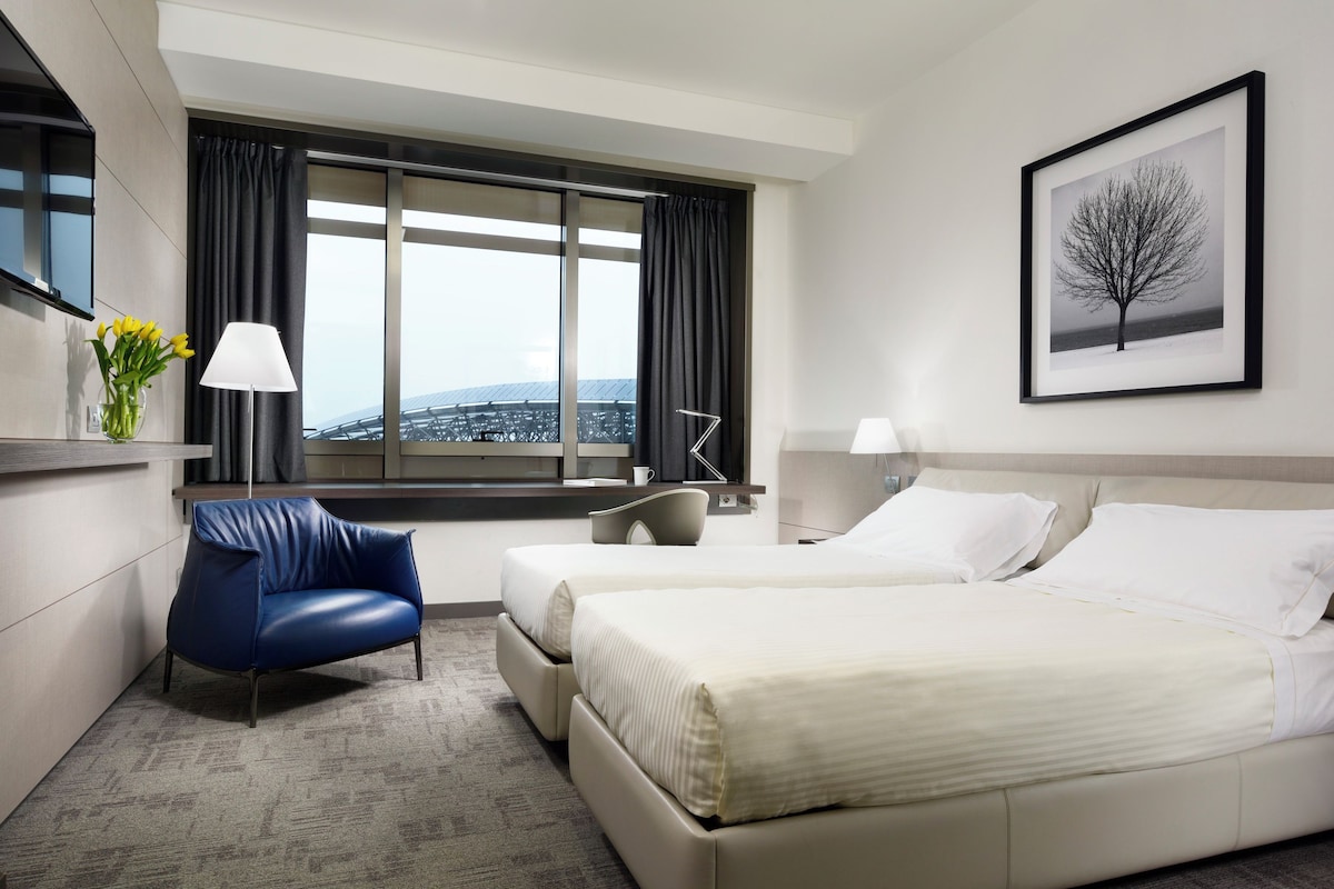A hotel room with a large bed, a chair, a desk, a window with curtains, a framed picture on the wall, and a vase with flowers on the desk.