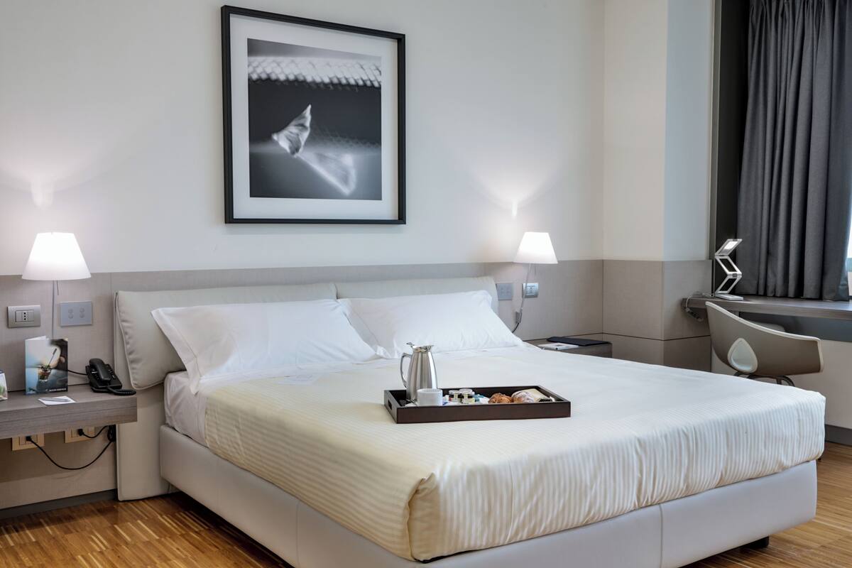 Une chambre d’hôtel avec un grand lit, des lampes de chevet, un tableau encadré au mur, un plateau avec des articles pour le petit-déjeuner et un bureau avec un téléphone et une lampe.
