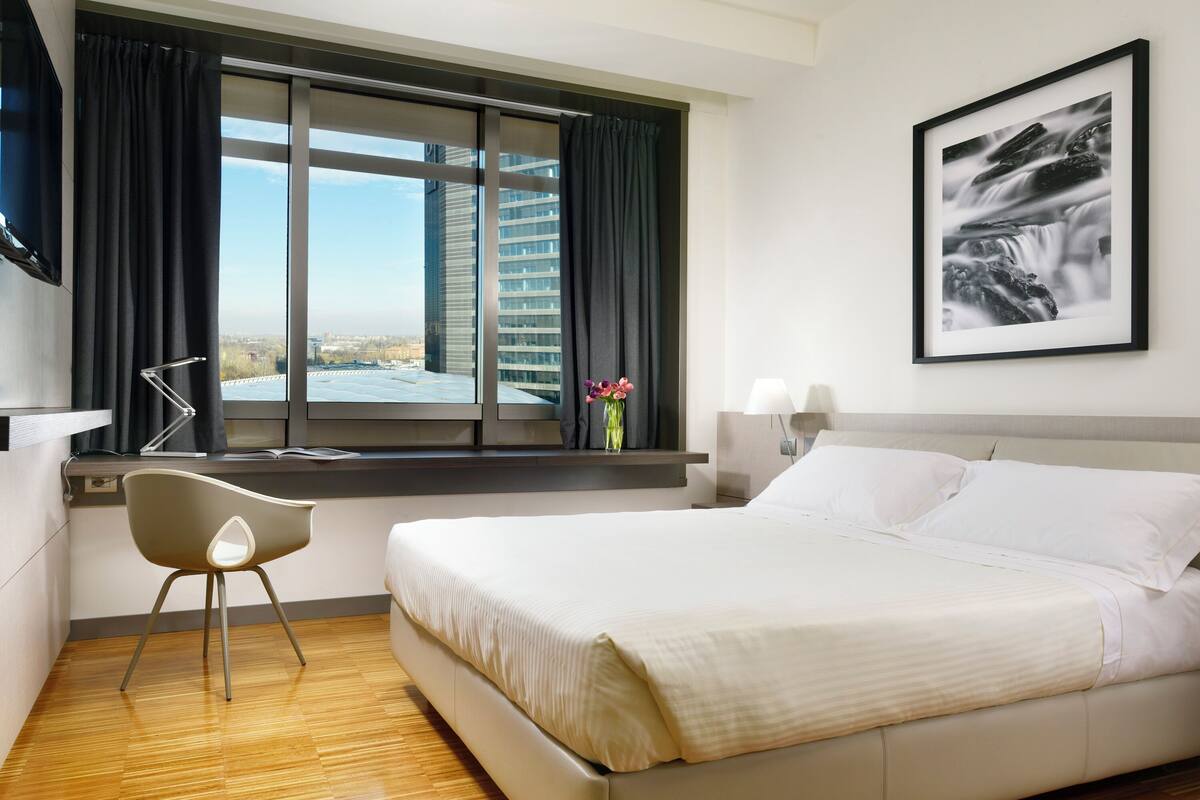Une chambre d’hôtel avec un lit, un bureau, une chaise, une fenêtre avec des rideaux, un tableau encadré au mur et une vue sur les bâtiments à l’extérieur.