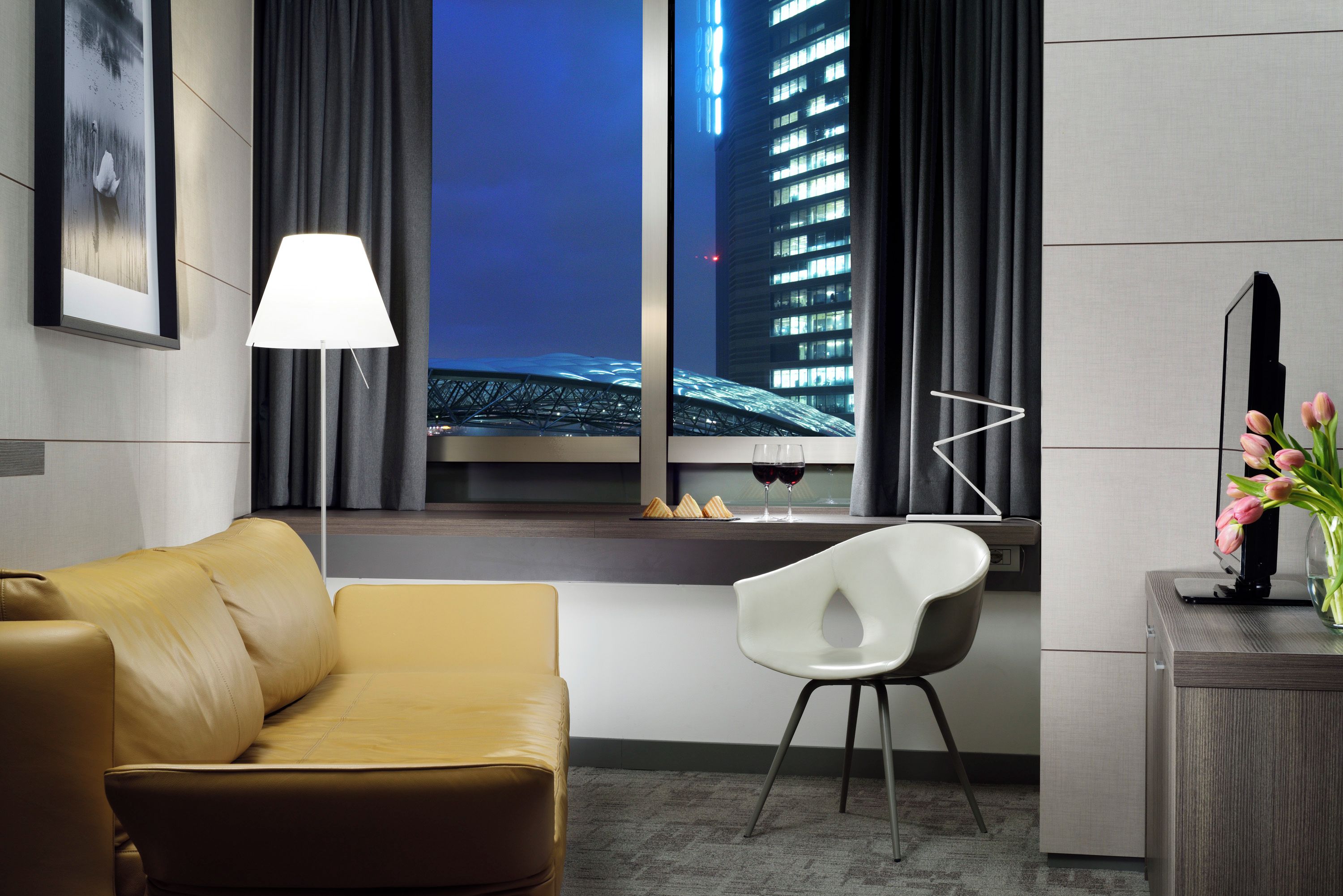 A modern hotel room with a leather sofa, a white chair, a desk with a vase of flowers, a lamp, and a view of a cityscape through a window.