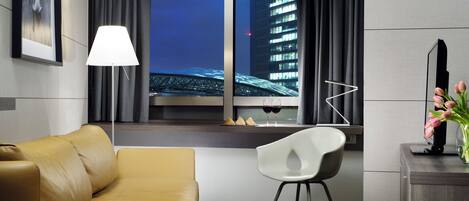 A modern hotel room with a leather sofa, a white chair, a desk with a vase of flowers, a lamp, and a view of a cityscape through a window.