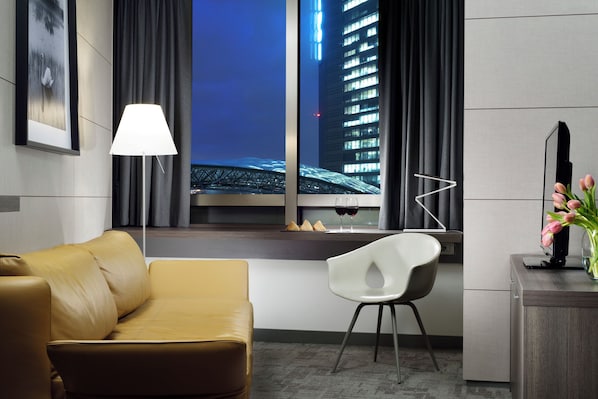 Ein modernes Hotelzimmer mit einer Ledersofa, einem weißen Sessel, einem Schreibtisch mit Blumenvase, einer Lampe und Blick auf eine Stadtlandschaft durch ein Fenster.