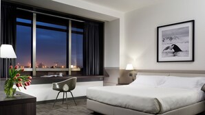 A modern hotel room with a large bed, a desk, a chair, a window with curtains, a framed picture on the wall, and a vase with flowers on the desk. - UNA Hotels San Vitale Bologna (Bologna)