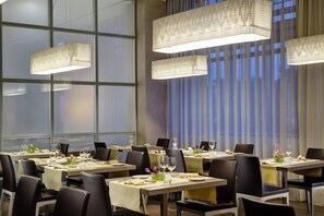 Un comedor con varias mesas dispuestas para comidas, ventanas amplias y lámparas colgantes.