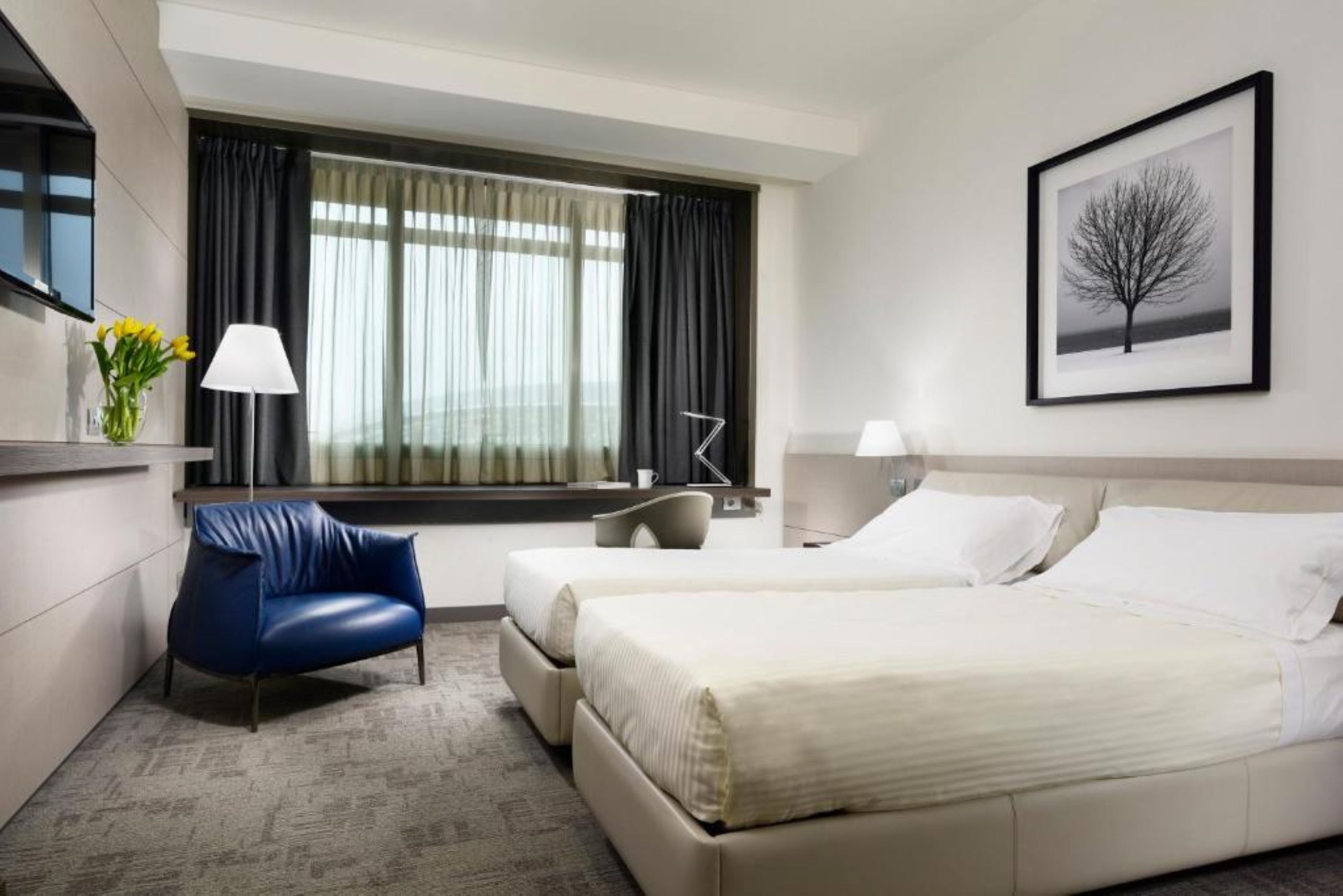 Hotellihuone, jossa on sänky, tuoli, työpöytä, lamppu, taulu seinällä ja ikkuna, jossa on verhot.