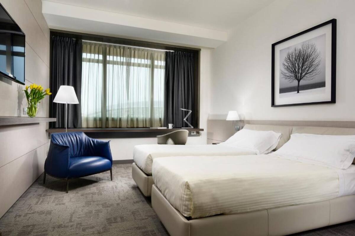 Une chambre d’hôtel avec un lit, une chaise, un bureau, une lampe et une fenêtre avec des rideaux.