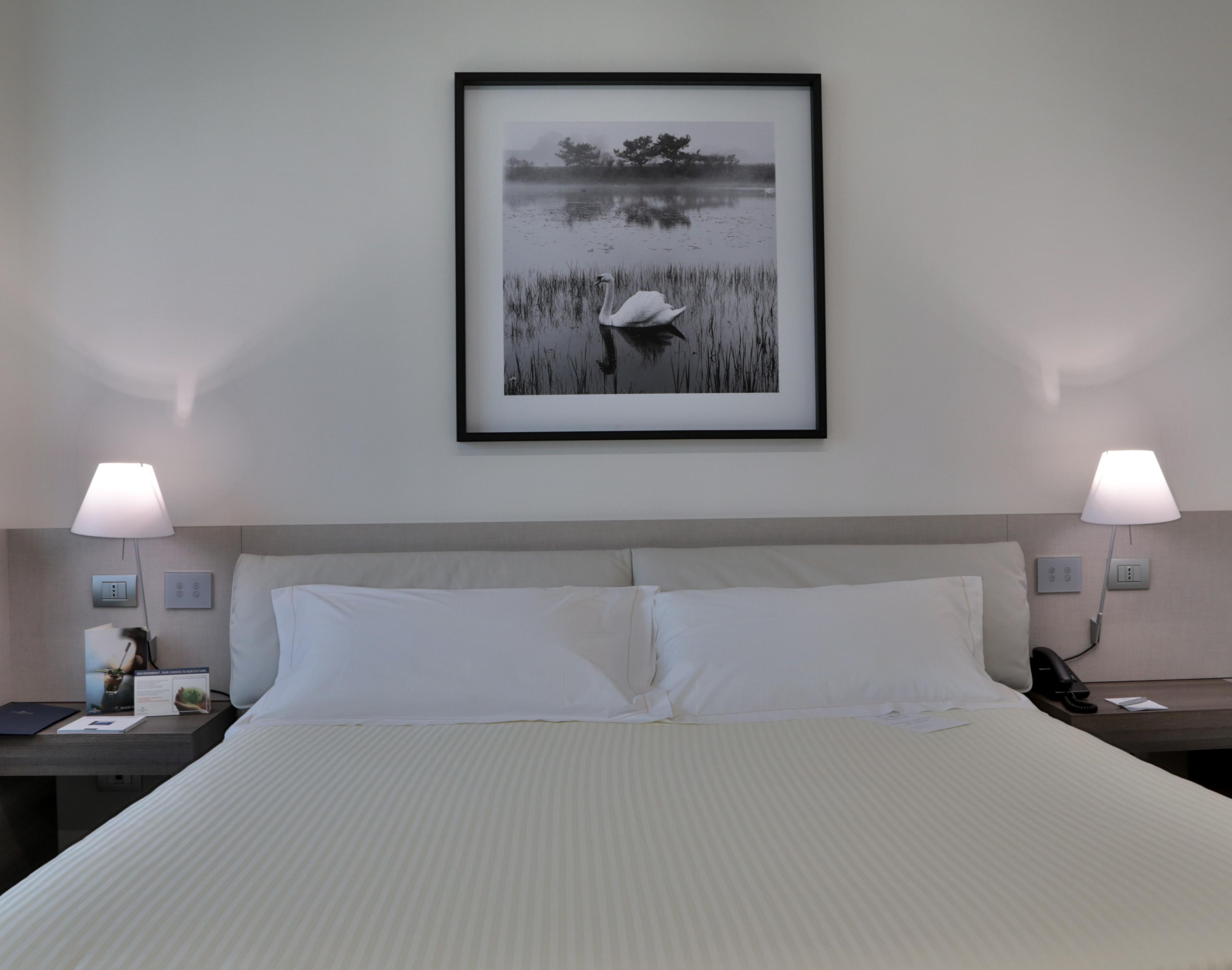 A hotel room with a large bed, two bedside lamps, a framed picture of a bird and water, and a nightstand with a lamp and a phone.