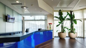 A reception desk with a blue finish, a flat-screen TV mounted on the wall, and two potted plants with large green leaves. - UNA Hotels San Vitale Bologna (Bologna)
