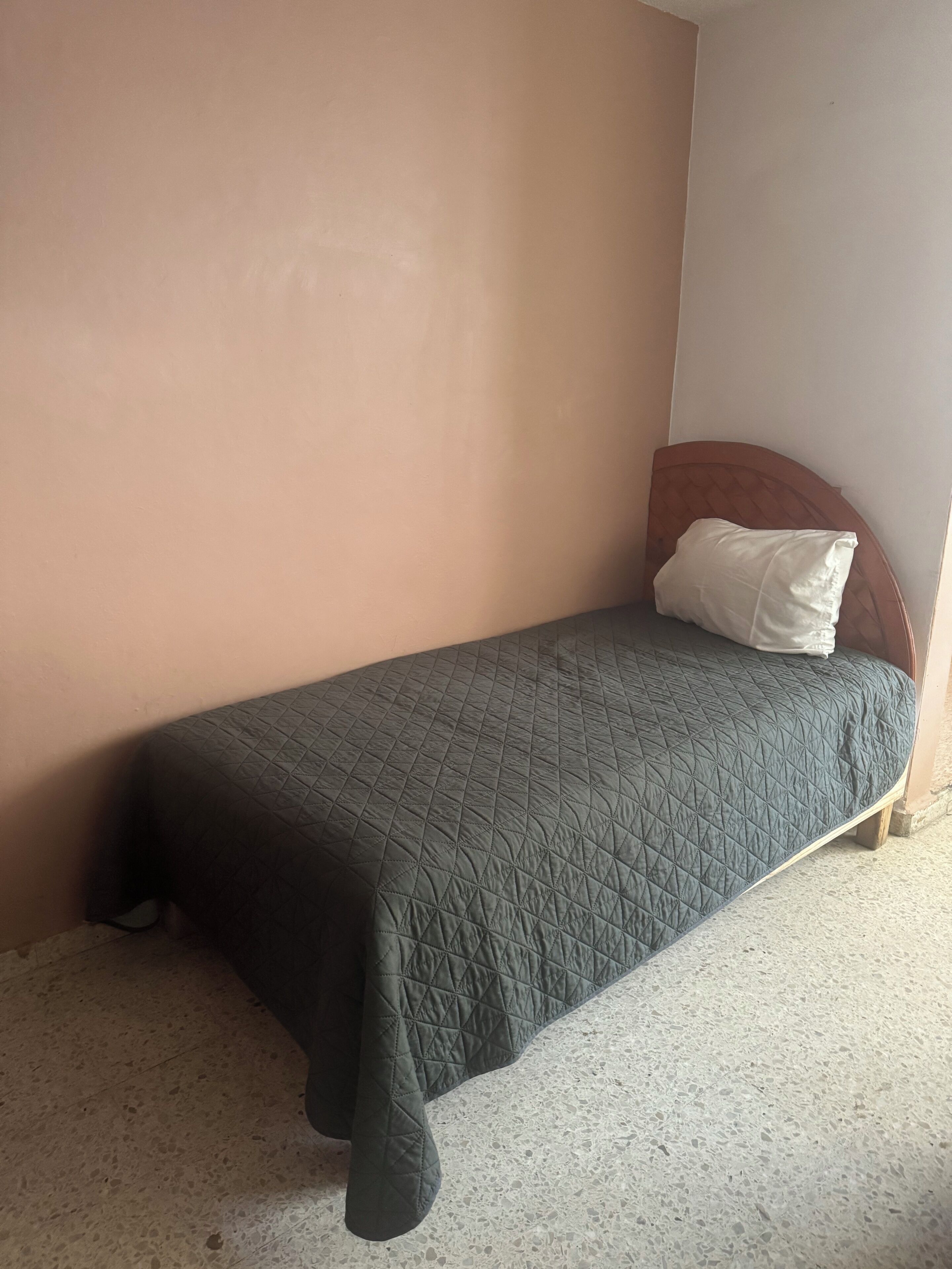 Un lit simple avec une couverture matelassée de couleur sombre, une tête de lit en bois et un oreiller blanc. La pièce est dotée d’un mur de couleur beige unie et d’un sol à motifs.