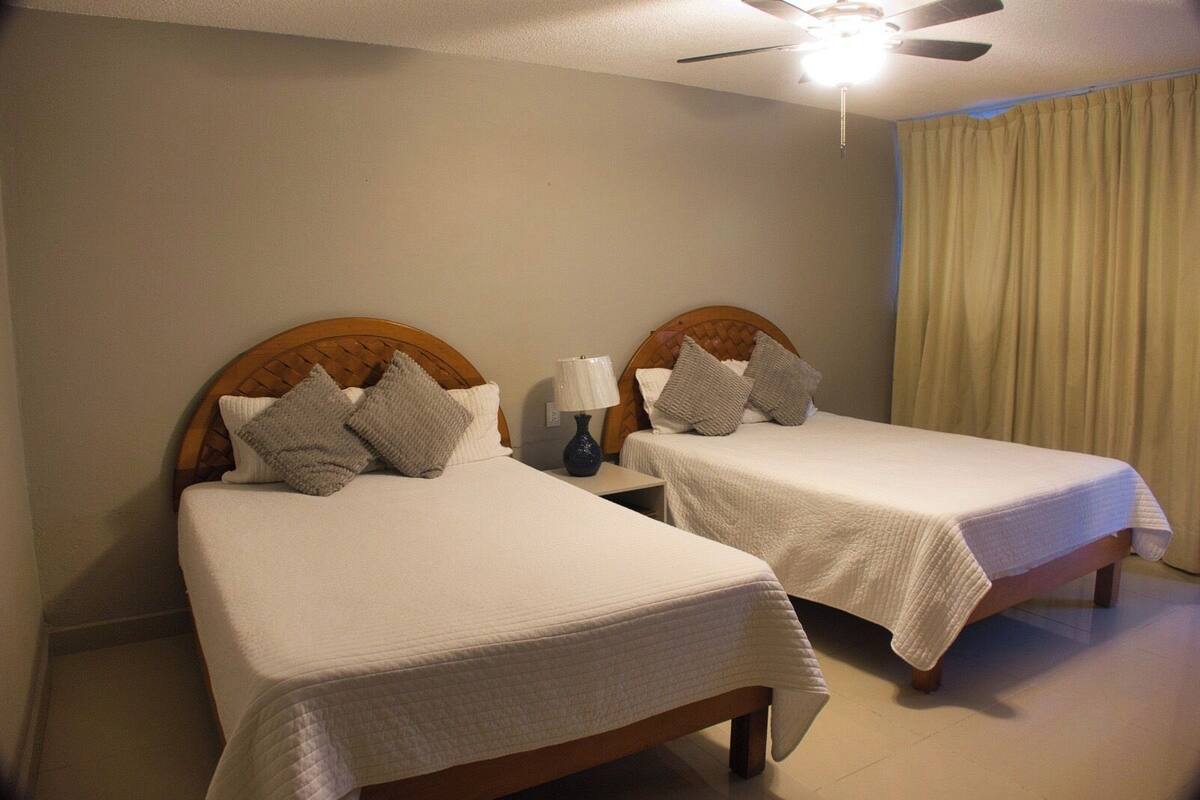 A hotel room with two single beds, a ceiling fan, a nightstand with a lamp, and a window with curtains.
