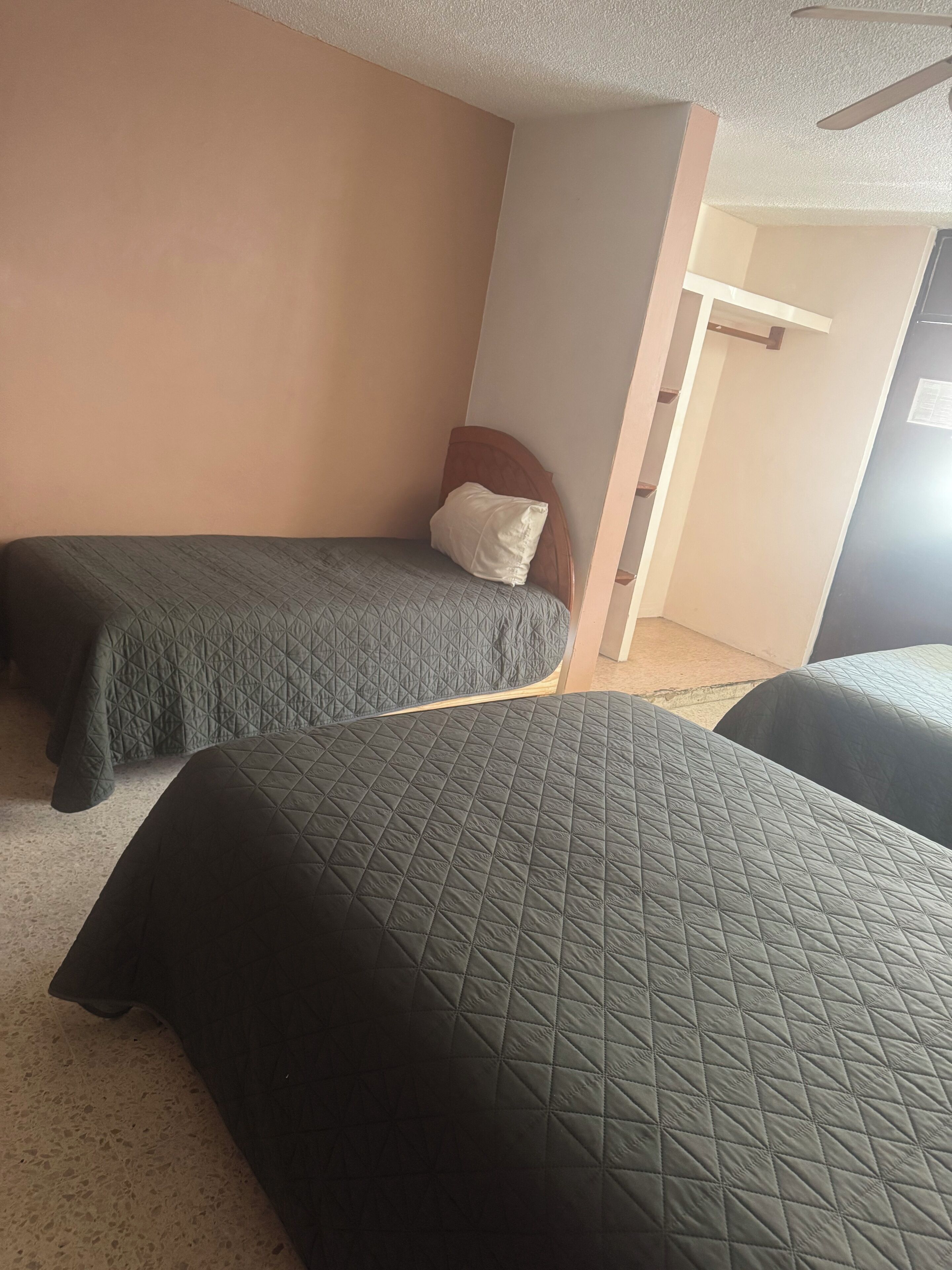 Een kamer met twee bedden, elk met een gewatteerde spreideken. Het een bed heeft een houten hoofdbord, het andere een wit kussen. De muren zijn beige en er is een deur naar een andere kamer.