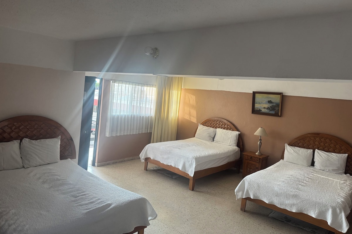 A hotel room with three beds, a window with curtains, a painting on the wall, and a nightstand with a lamp.