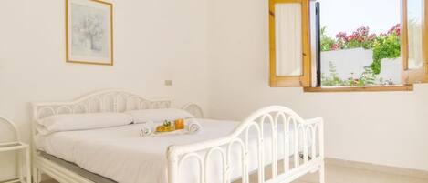 A white metal bed with a white mattress and pillows, a bedside table, a framed picture on the wall, and a window with a view of greenery and flowers.
