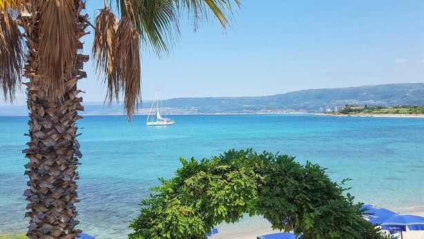 Une plage avec une eau d’un bleu limpide, un voilier et un palmier.