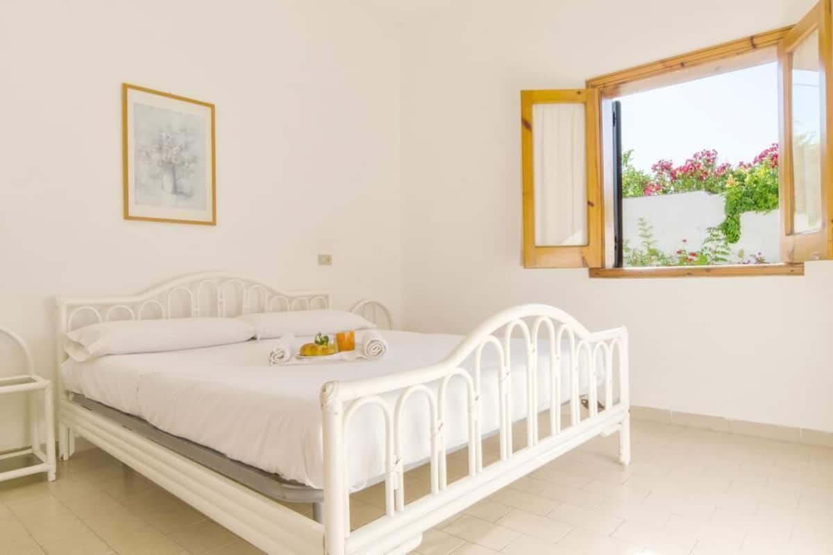 A white metal bed with a white mattress and pillows, a bedside table, a framed picture on the wall, and a window with a view of greenery and flowers.