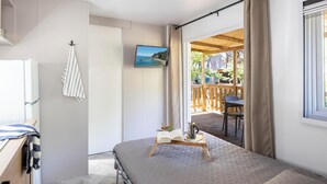 Un dormitorio con cama, televisión, ventana con cortinas, mesa de madera con un libro y una cafetera, y vista a una terraza exterior.