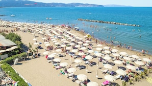 Una playa con numerosos sombrillas y tumbonas, un rompeolas rocoso y barcos en el agua.