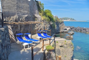 Una terraza frente al mar con tumbonas azules, un muro de piedra y una costa rocosa.