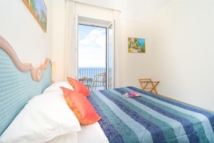 Un dormitorio con cama, almohadas, una silla, una mesita y una puerta al balcón.