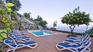 Ein Poolbereich mit Liegestühlen rund um einen rechteckigen Pool, eine Steinmauer und Meerblick.