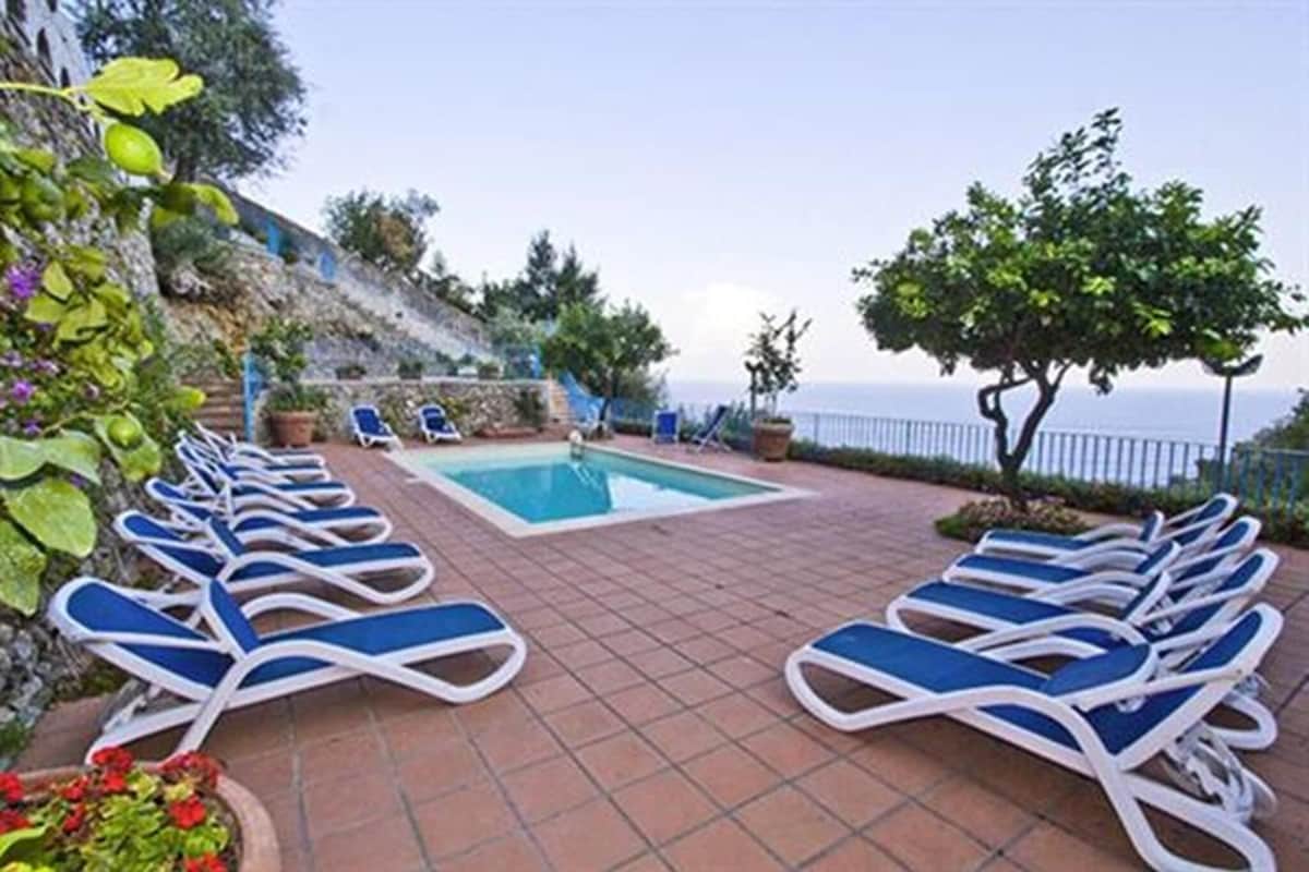 A poolside area with lounge chairs arranged around a rectangular pool, a stone wall, and a view of the sea.