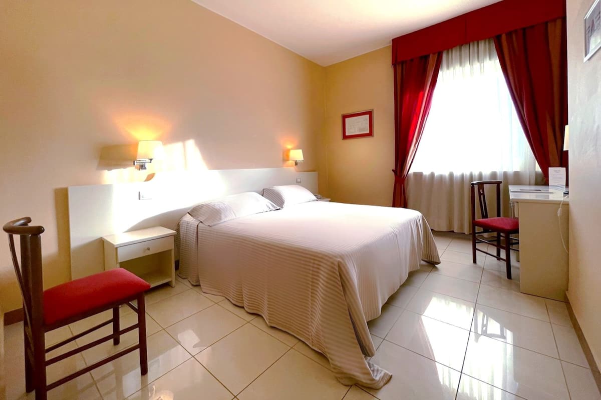 A hotel room with a bed, bedside tables, a desk, a chair, and a window with curtains.