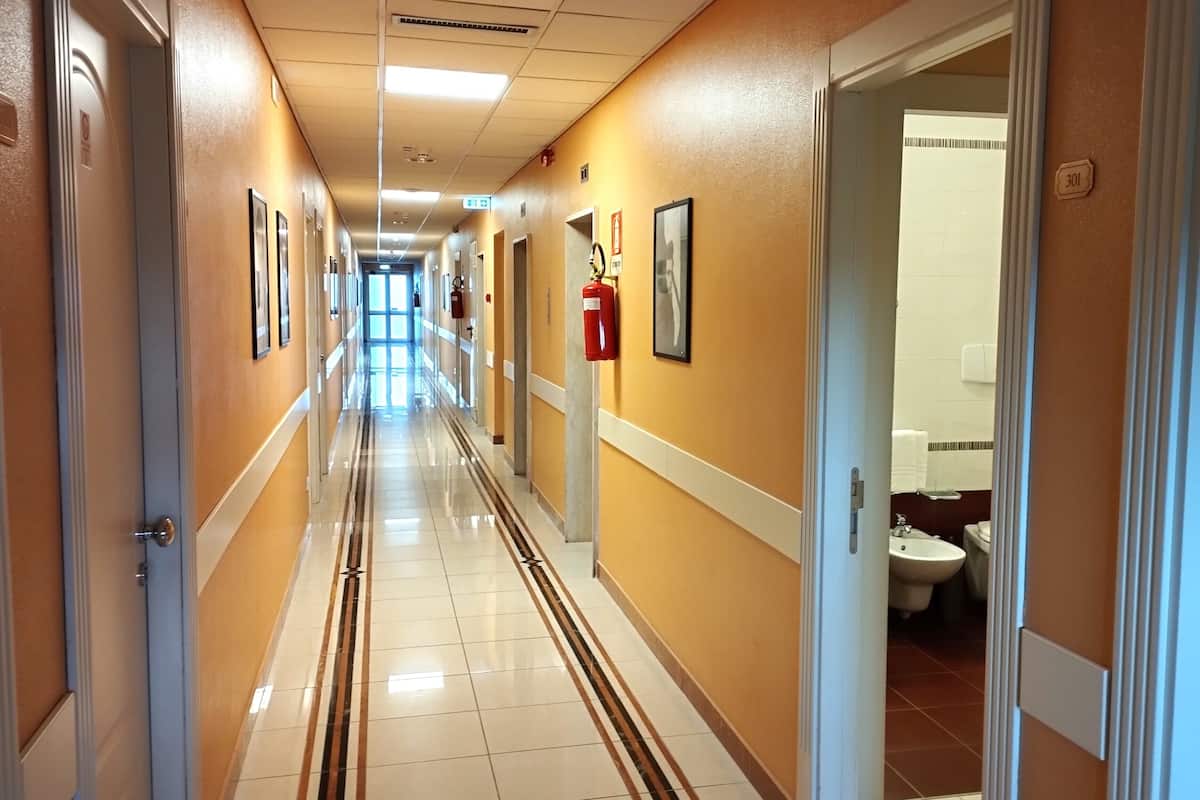 A hotel hallway with a tiled floor, doors on both sides, framed pictures on the walls, and a visible bathroom with a toilet and sink.