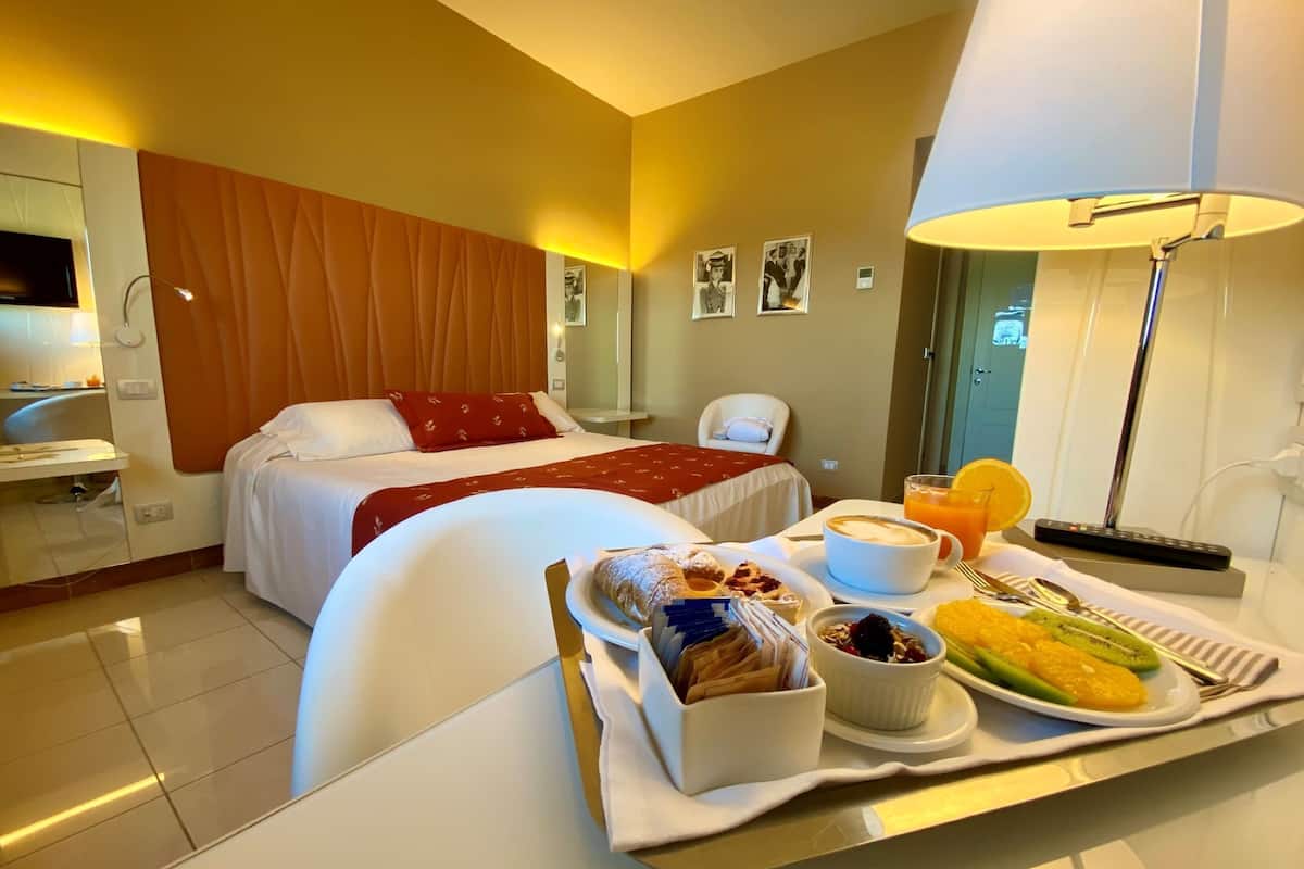 A hotel room with a bed, a desk with a tray of breakfast, a chair, and a lamp.