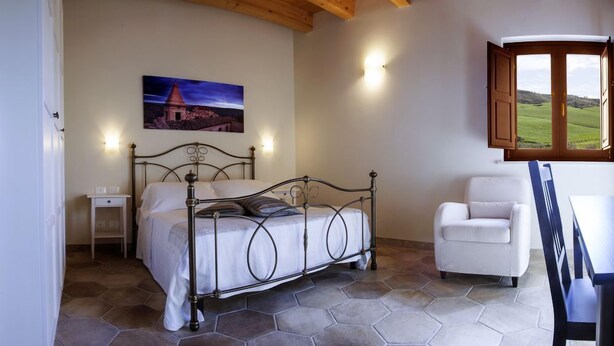 A bedroom with a metal bed frame, a white chair, and a view of a green landscape through a window. - Arucimeli Resort (Ragusa)
