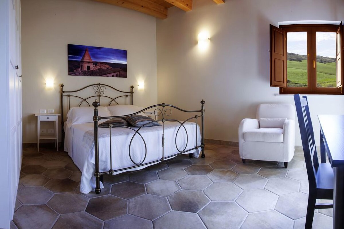 A bedroom with a metal bed frame, a white chair, and a view of a green landscape through a window.
