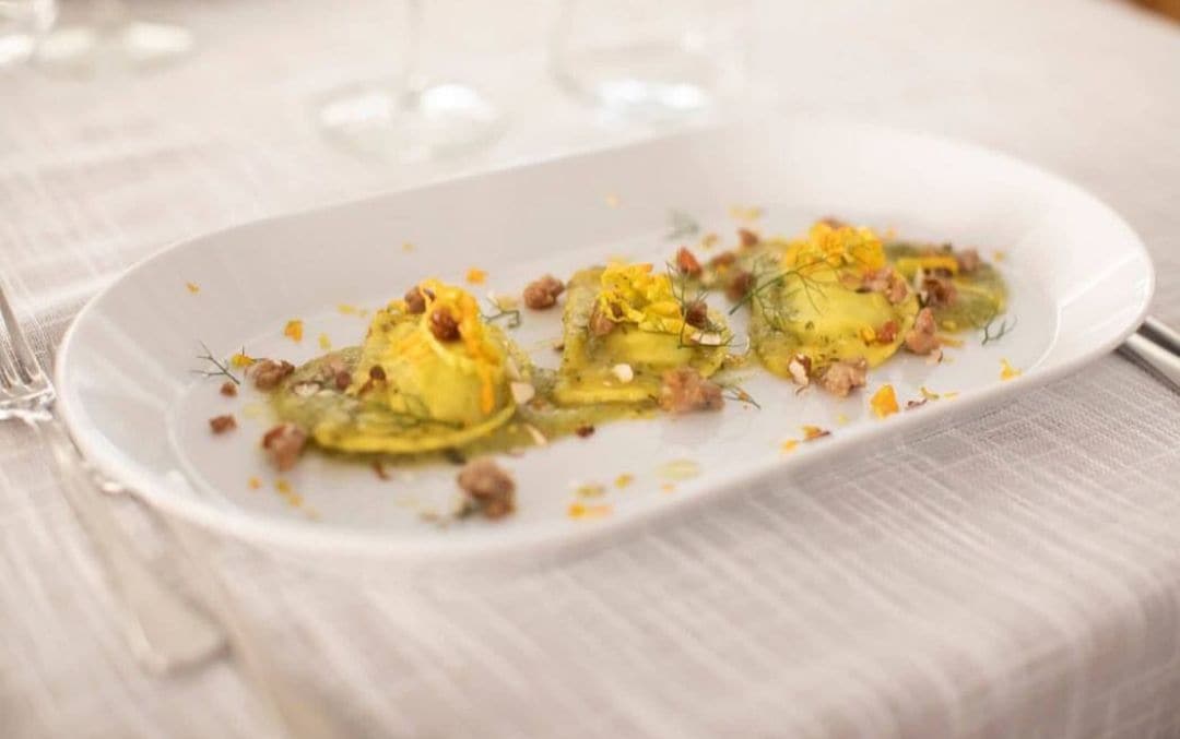 Une assiette blanche contenant trois raviolis, décorés d’herbes et de petits morceaux de viande, est posée sur une table recouverte d’une nappe blanche.