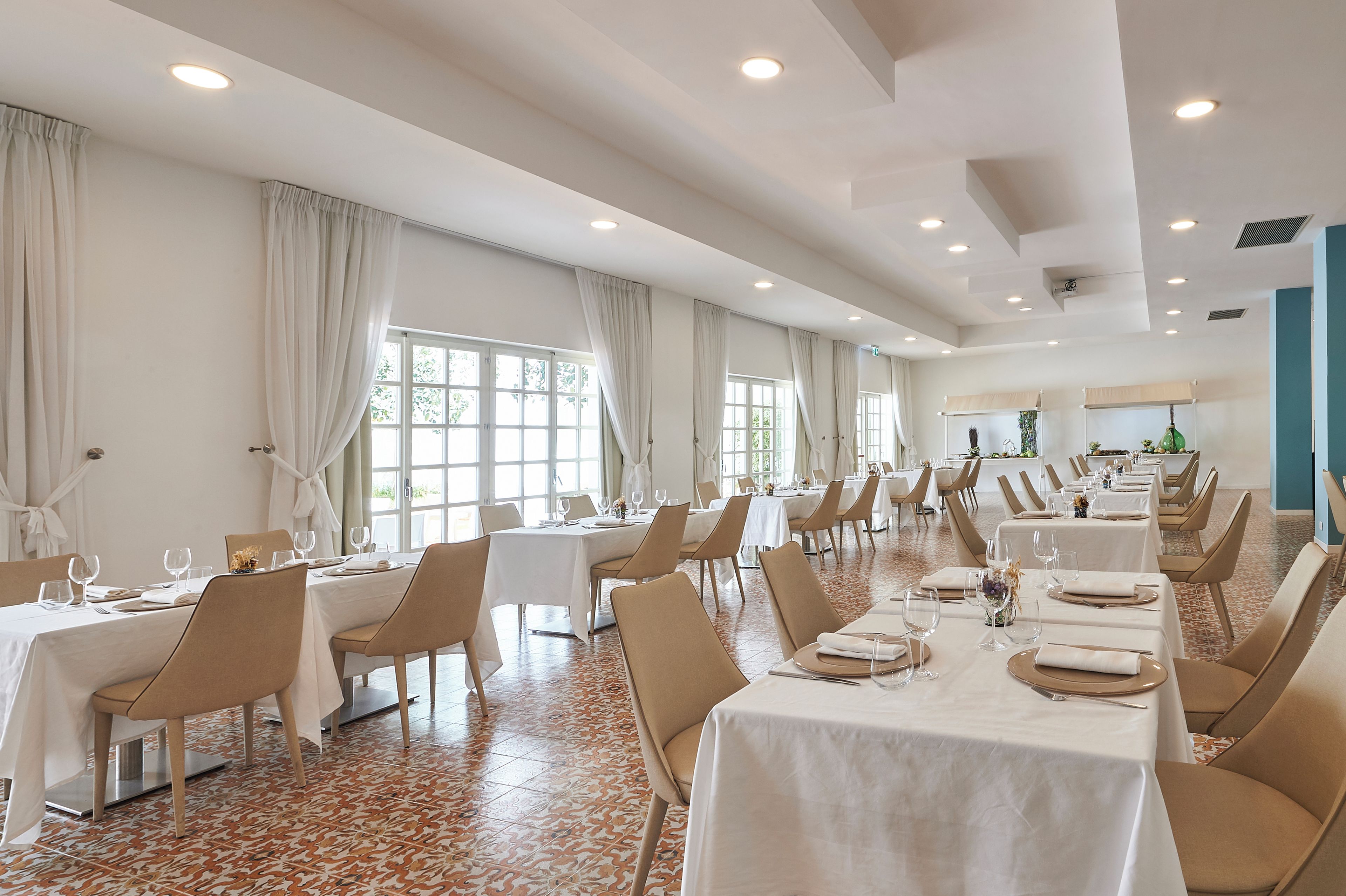A spacious dining area with multiple tables and chairs, large windows with curtains, and a tiled floor.