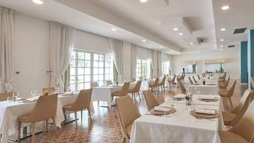 Un'ampia sala da pranzo con diversi tavoli e sedie, grandi finestre con tende e un pavimento piastrellato.