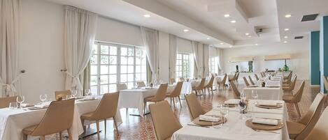 A spacious dining area with multiple tables and chairs, large windows with curtains, and a tiled floor.