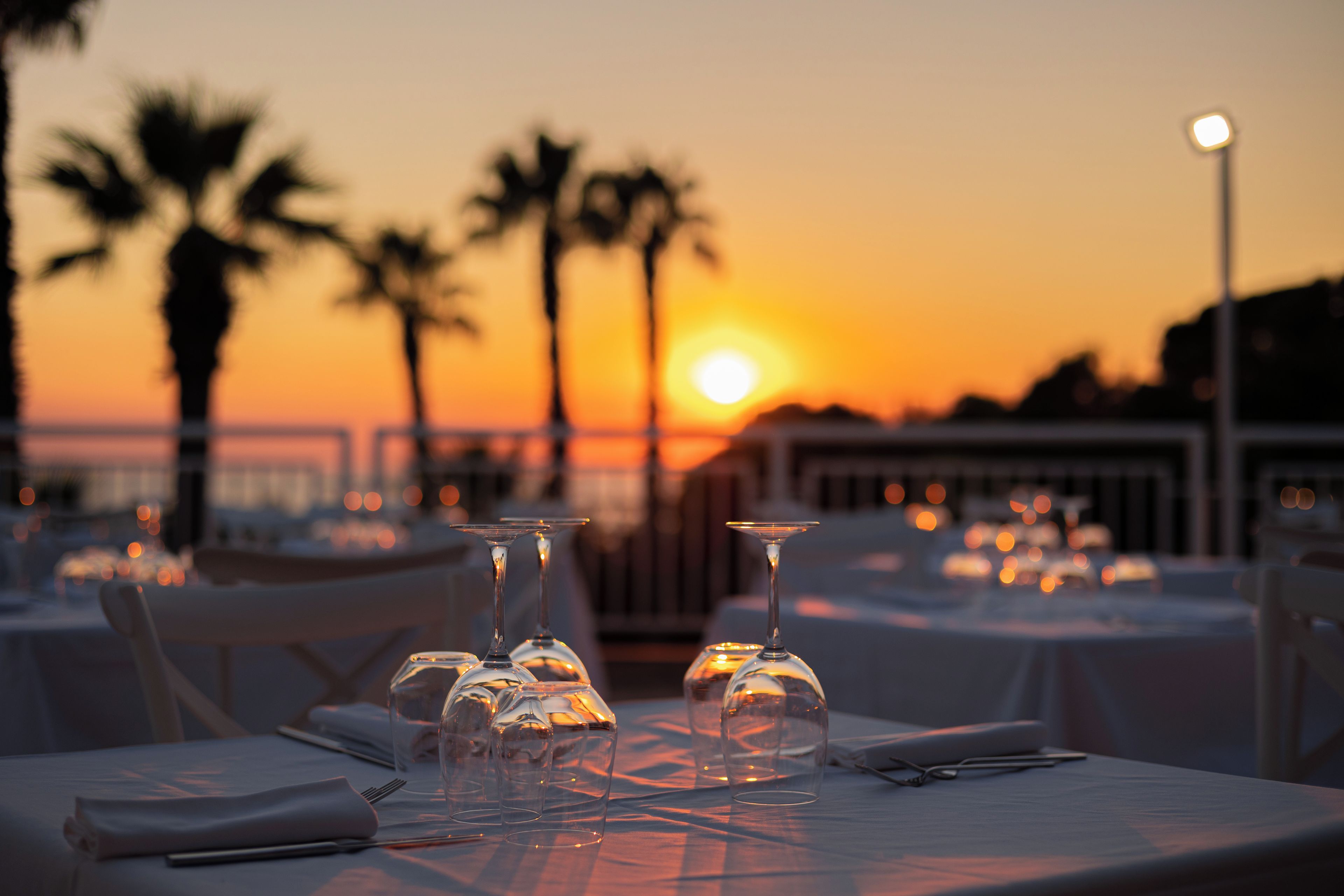 Tavola imbandita per la cena, con calici di vetro trasparenti e un tovagliolo piegato, affacciata su un tramonto con palme sullo sfondo.
