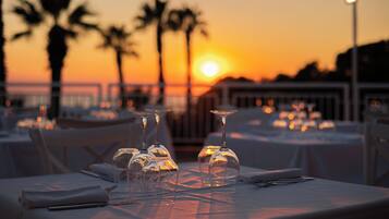 Tavola imbandita per la cena, con calici di vetro trasparenti e un tovagliolo piegato, affacciata su un tramonto con palme sullo sfondo.