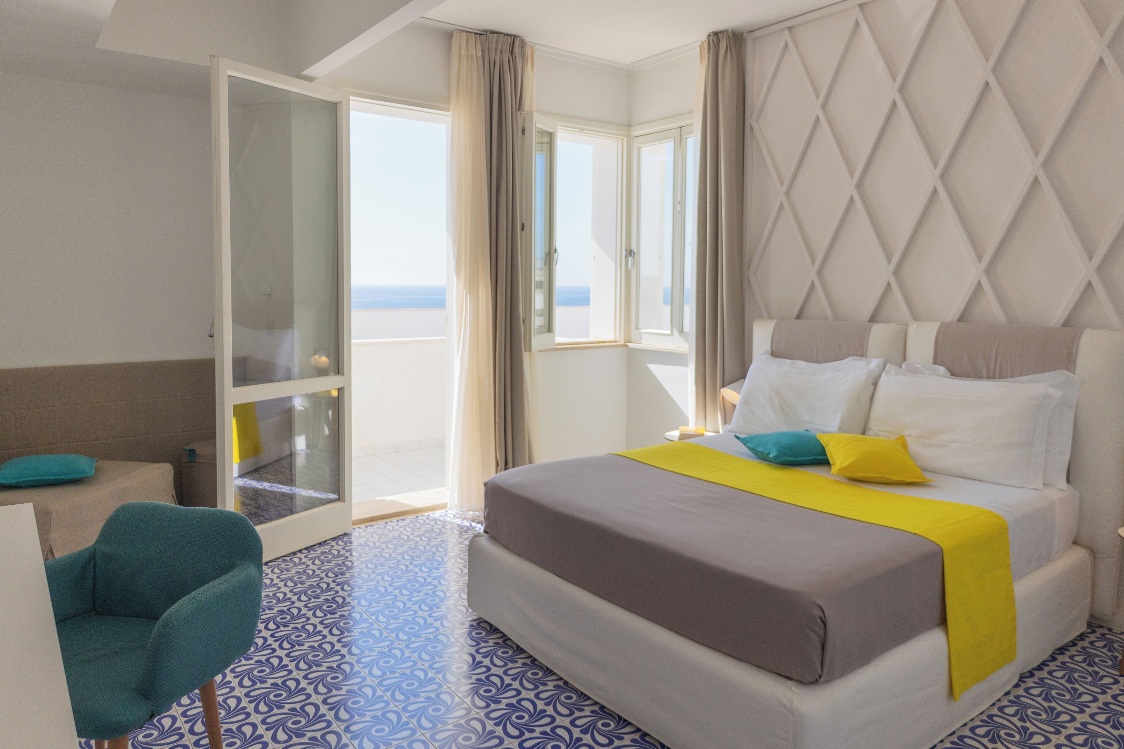 A hotel room with a bed, a chair, a desk, and a view of the sea through large windows.