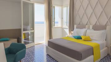 A hotel room with a bed, a chair, a desk, and a view of the sea through large windows.