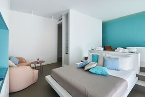 Une chambre d’hôtel moderne, équipée d’un lit, d’un canapé, d’une petite table avec un vase, et d’une salle de bain avec un lavabo et un miroir blancs.