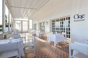 Un espace de restauration intérieur avec des tables dressées, un sol carrelé et une vue dégagée sur l’extérieur grâce à de grandes fenêtres.