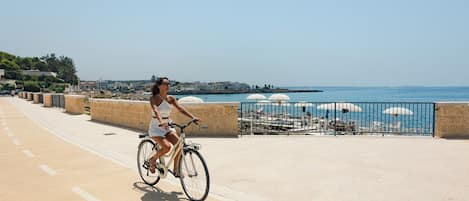 Una mujer pedaleando en bicicleta por un camino pavimentado al lado de un paseo marítimo, con vista al mar y edificios al fondo.