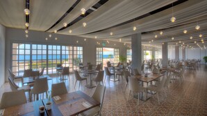 A spacious dining area with multiple tables and chairs, large windows offering a view of the sea, and hanging spherical light fixtures. - Grand Hotel Riviera CDS Hotels (Nardò)
