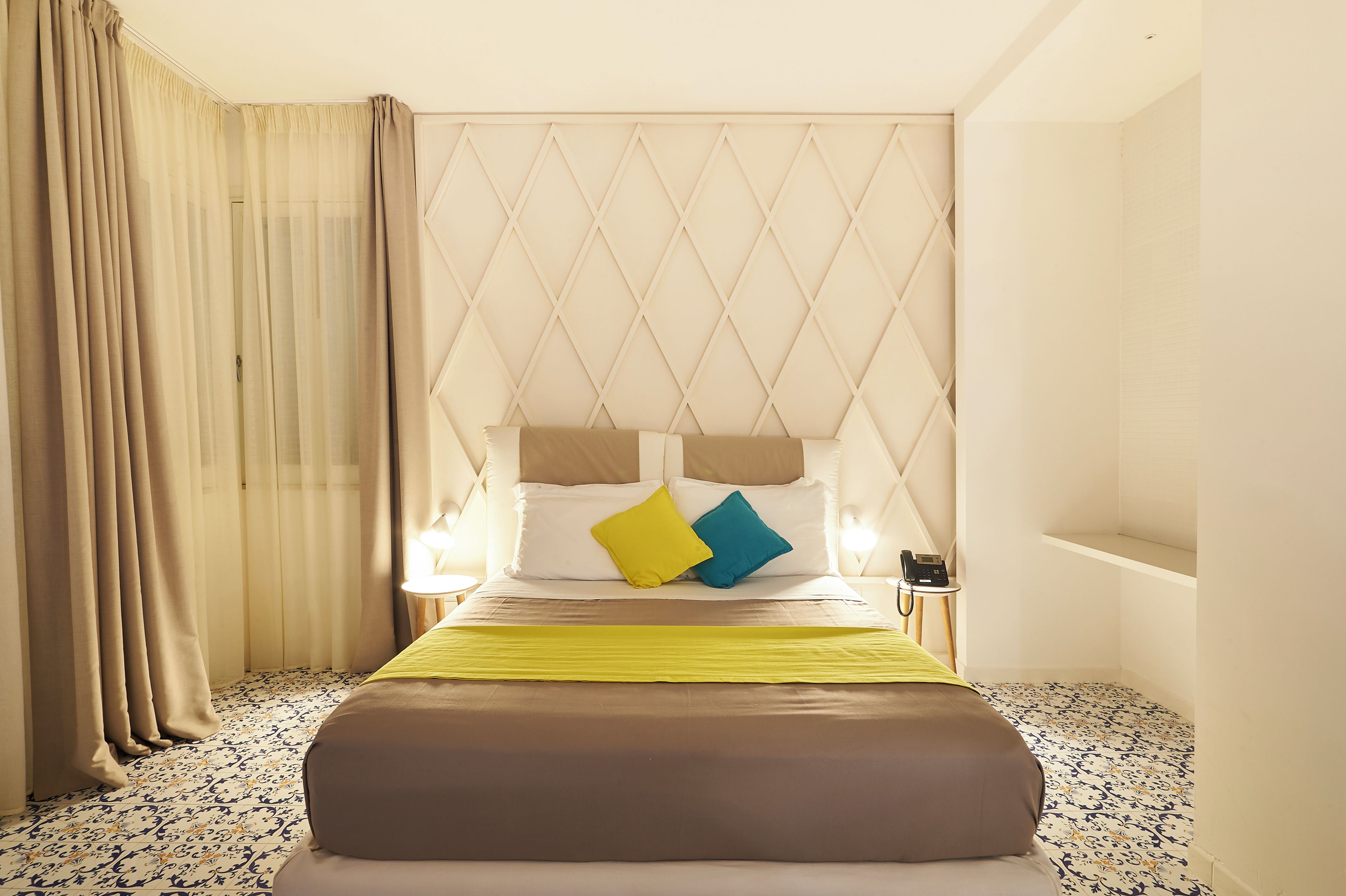 Una habitación de hotel con cama, mesita de noche, lámpara y ventana con cortinas.