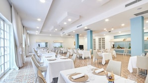Un grand espace dédié à la salle à manger, avec plusieurs tables dressées, de grandes fenêtres d’un côté et un coin cuisine à l’arrière-plan.