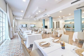 A spacious dining area with multiple tables set for dining, large windows on one side, and a kitchen area in the background. - Grand Hotel Riviera CDS Hotels (Nardò)